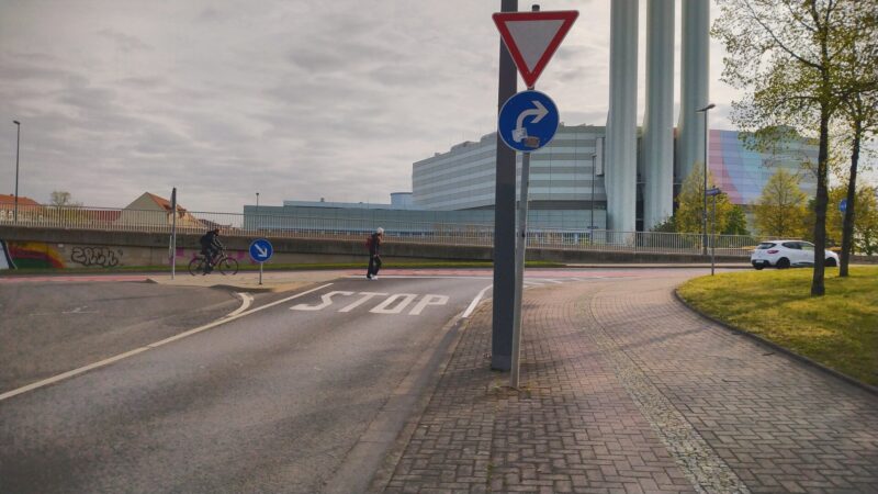 Eine Straße mit großer, weißer Aufschrift "STOP". Im Vordergrund ein Verkehrsschild mit Rechts-Abbieger und Vorfahrt Achten.