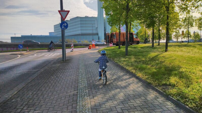 Rad- und Fußweg rechts neben der Straße zum Kraftwerk Nossener Brücke. Auf der Straße steht "STOP", ein Vorfahrt-Achten-Schild mit Rechtsabbieger. Im Hintergrund sieht man Bauarbeiter. Am Wagen lehnt ein rotes STOP-Schild.