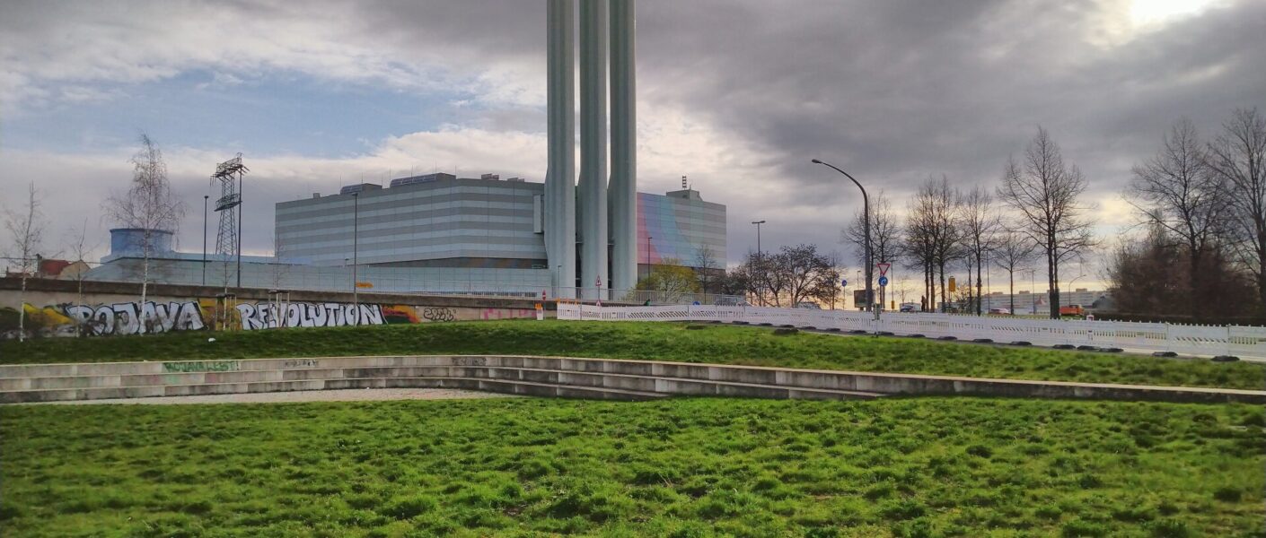 Im Vordergrund der Ebertplatz mit großer, grober Wiese. Im Hintergrund das Kraftwerk Nossener Brücke mit den drei Schornsteinen.