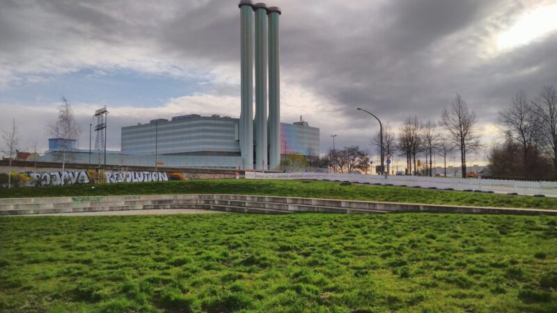 Im Vordergrund der Ebertplatz mit großer, grober Wiese. Im Hintergrund das Kraftwerk Nossener Brücke mit den drei Schornsteinen.