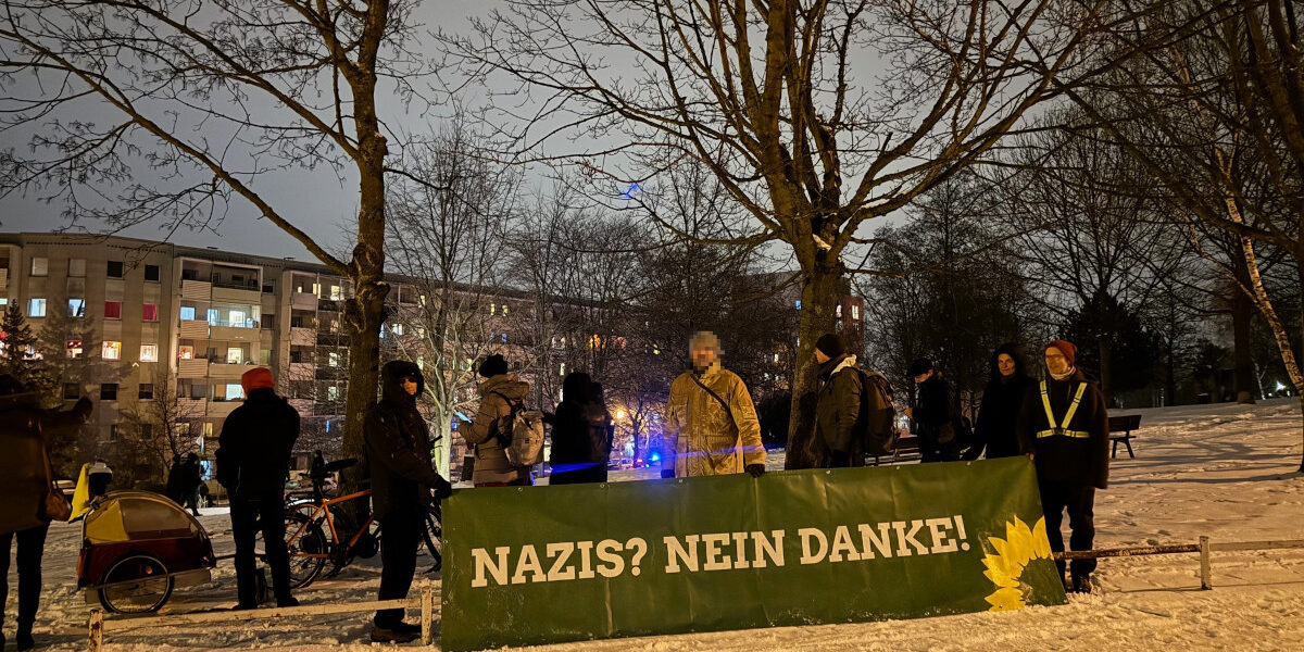 Ein grünes Banner mit Sonnenblume und der Aufschrift "Nazis nein Danke" wird gehalten von einer kleinen Menge Menschen. Es ist dunkel, es liegt Schnee. Im Hintergrund die Neubaublöcke von Gorbitz und zwei Bäume.