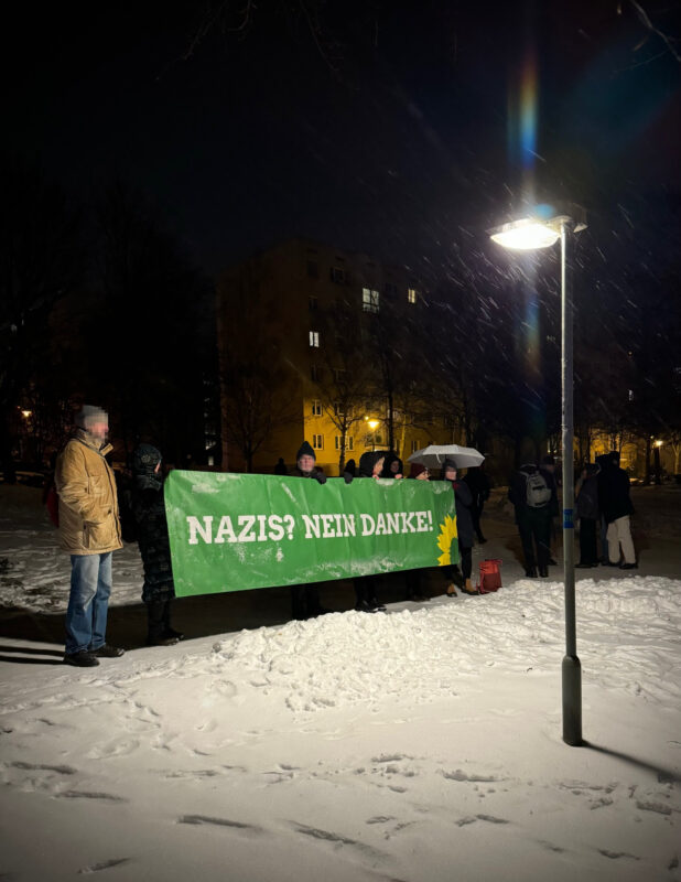 Ein grünes Banner mit Sonnenblume und der Aufschrift "Nazis nein Danke" wird gehalten von einer kleinen Menge Menschen. Es ist dunkel, es liegt Schnee. Beleuchtet von einer Straßenlampe.