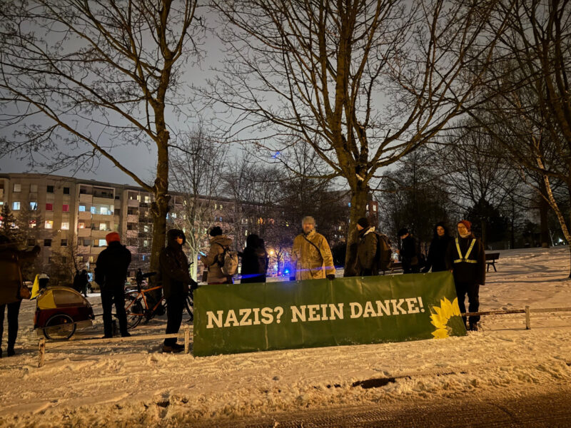 Ein grünes Banner mit Sonnenblume und der Aufschrift "Nazis nein Danke" wird gehalten von einer kleinen Menge Menschen. Es ist dunkel, es liegt Schnee. Im Hintergrund die Neubaublöcke von Gorbitz und zwei Bäume.