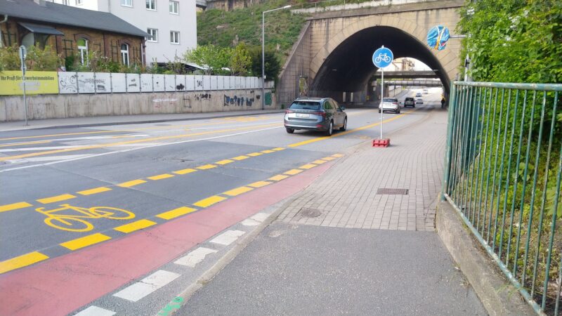 Straße mit gelber Radweg-Markierung. Die Straße führt unter einem Bahntunnel durch.