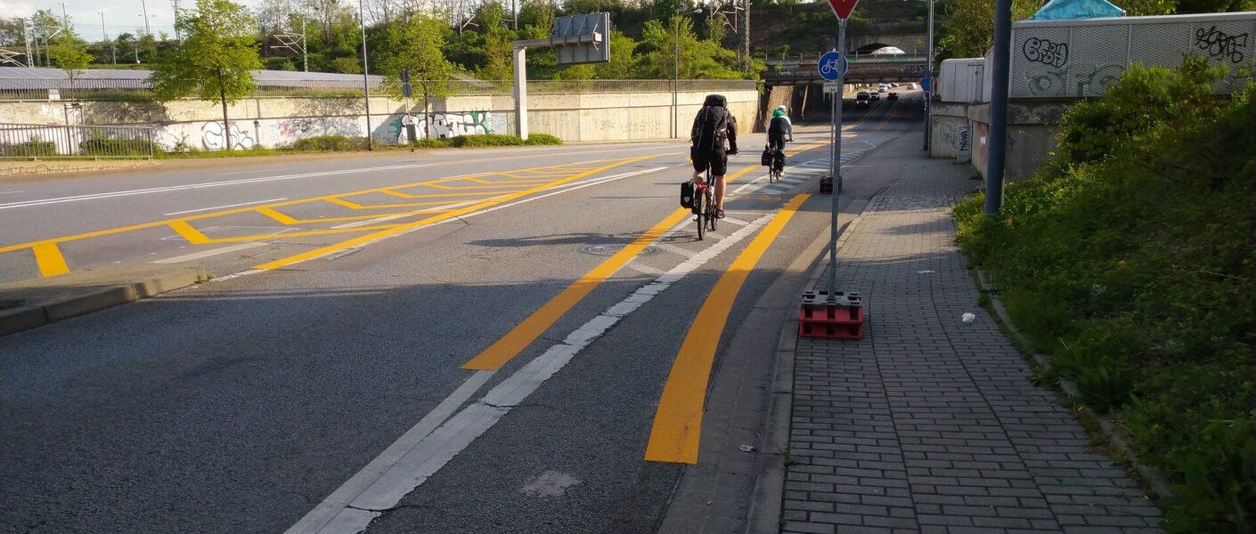 Straße mit vielen gelben Markierungen, zwei Radfahrer von hinten, keine Autos
