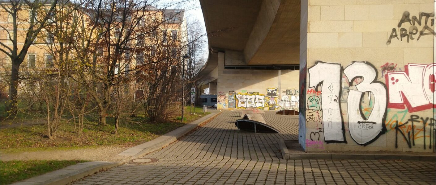 Im Vordergrund die Betongittersteine, rechts ein angsprühter Brückenpfeiler. Die Brücke überragt die befestigte Fläche. Links Büsche und Bäume ohne Laub. Dahinter links Löbtauer Mehrfamilienhäuser.