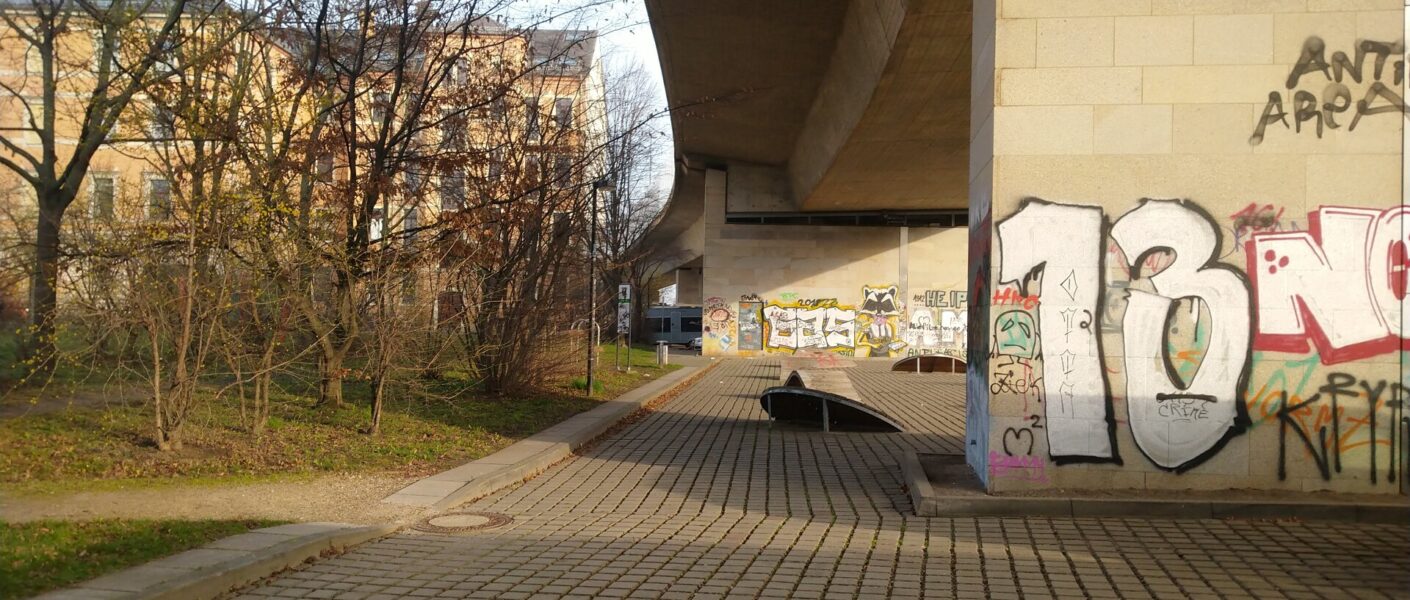 Im Vordergrund die Betongittersteine, rechts ein angsprühter Brückenpfeiler. Die Brücke überragt die befestigte Fläche. Links Büsche und Bäume ohne Laub. Dahinter links Löbtauer Mehrfamilienhäuser.