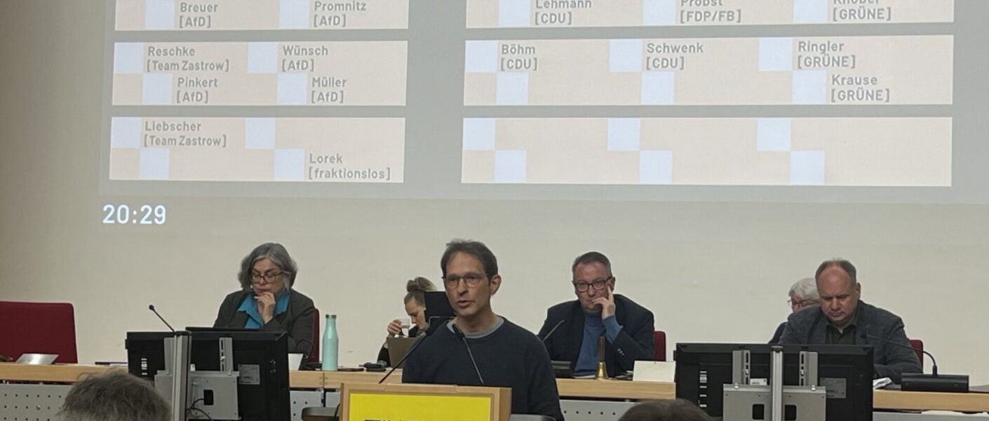Mensch steht am Rednerpult mit Dresden-Logo. Dahinter sitzen drei Personen.