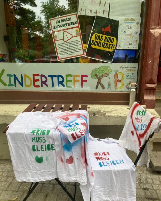 Vor dem Schaufenster steht ein Gartentisch und ein Stuhl. Darauf weiße T-Shirts mit bunter Schrift "#KIBO MUSS BLEIBEN"