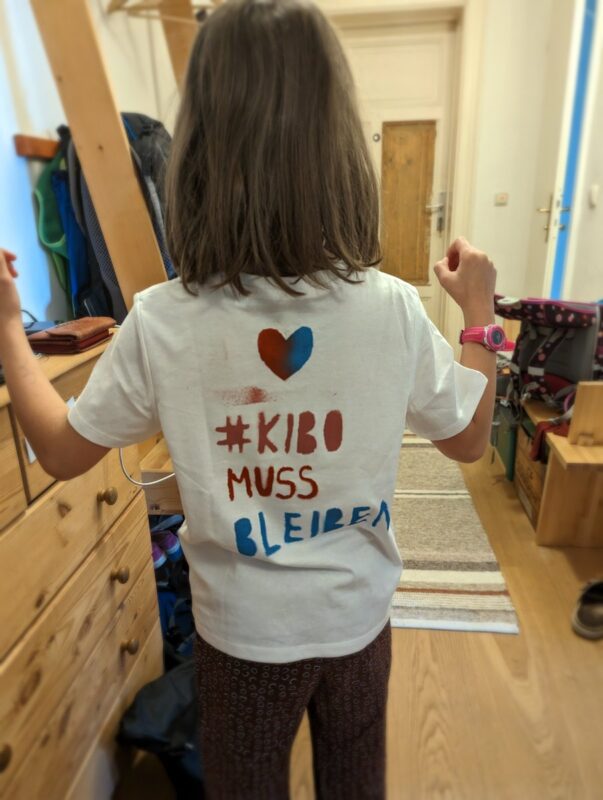 Ein Kind mit einem Demo-T-Shirt von hinten fotografiert. Auf dem Shirt steht "#KIBO MUSS BLEIBEN"