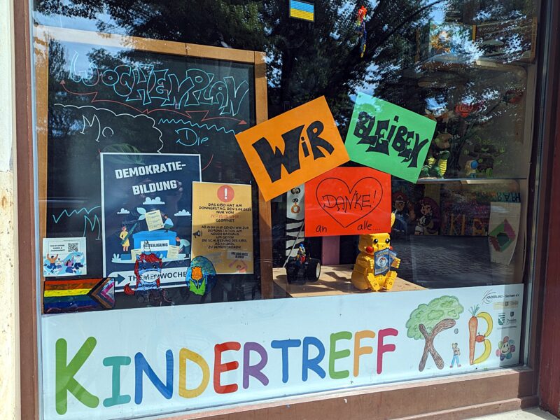 Foto des Schaufensters. Unten die bunten Buchstaben "Kindertreff KiBo". Mittig drei bunte A4-Zettel mit dem Text "Wir bleiben" und "Danke an alle" mit einem Herz.
