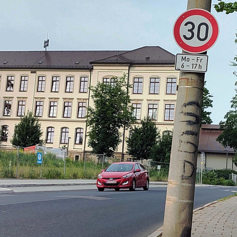 Ein Schulgebäude im Hintergrund. Davor eine Straße mit einem entgegenkommenden, roten Auto. Rechts ein dicker Beton-Mast mit einem Tempo 30 Schild und dem Zusatz Mo-Fr 10-17 Uhr.