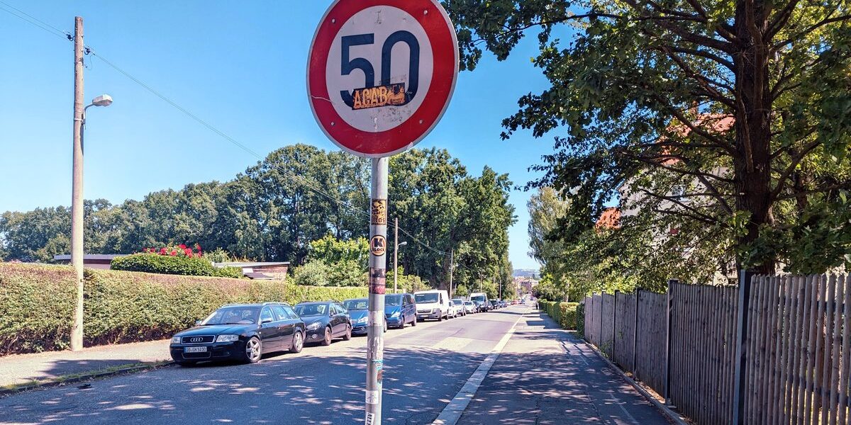 Eine Straße bergab fotografiert. Links paren Autos, rechts ein breiter Fußweg, der kaum von der Straße unterscheidbar ist. In Bildmitte ein Tempo-50 Schild ohne Zusatz am dünnen Metall-Mast