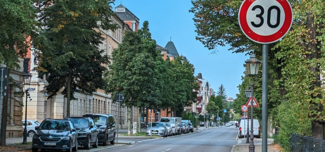 Im Vordergrund, rechts ein Tempo 30 Schild. In der Mitte eine leere Straße. Links und rechts sind Parkstreifen und Baumstreifen.