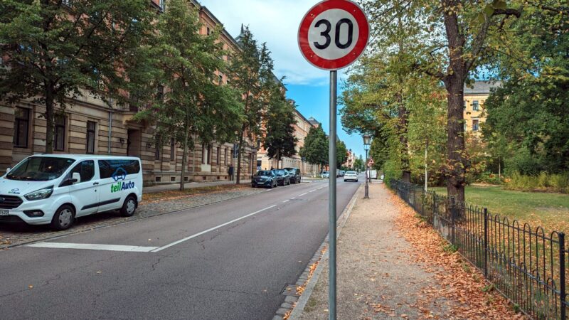 Eine Straße, kein Verkehr. Im Vordergrund ein Tempo 30-Schild. Links auf dem Parkstreifen ein weißer teilAuto Kleinbus.