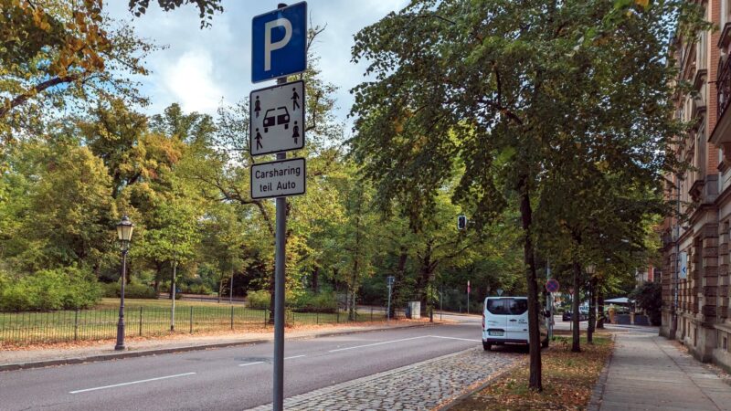Im Vordergrund ein Parkplatz-Schild. Darunter das Schild für Carsharing: mittig ein halbiertes Auto mit vier Menschen in den Ecken. Darunter ein Schild mit dem Text "Carsharing teil Auto". Auf dem Parkstreifen sind zwei leere Parkplätze und ganz vorne ein weißer Bus von teilAuto.