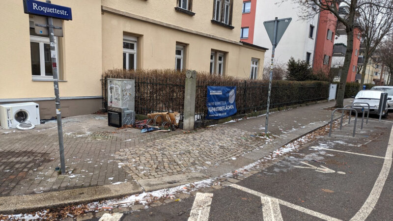 Eine Straßenecke, rechts sind drei neue Fahrradbügel, auf der Straße ist ein Fußgängerüberweg markiert. Direkt vor dem Haus liegt Müll (Herd, Waschmaschine)