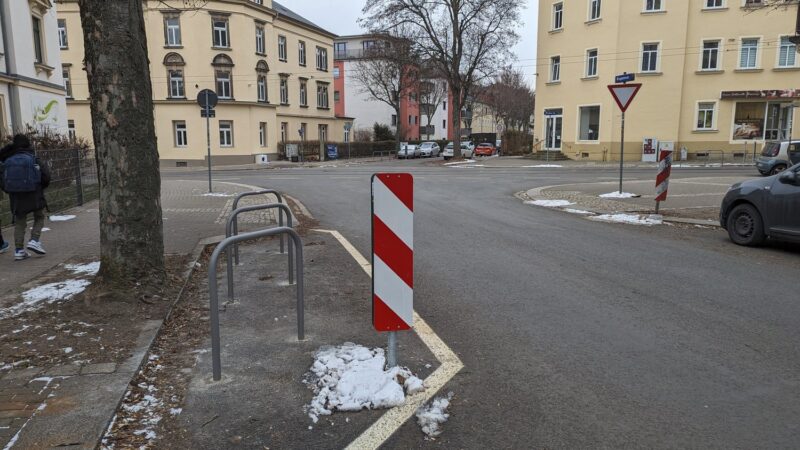 Drei Fahrradbügel schräg postioniert, davor eine Warnbarke., links davon ein Baum.