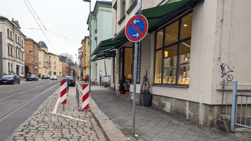 zwei Warnbarken vor drei Fahrradbügel, rechts ein Parken verbten (in beide Richtungen), rechts ein Ladengeschäft mit Markiesen.