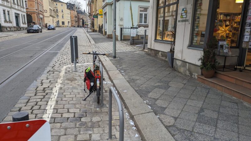 drei Fahrradbügel hintereinander. Am mittleren steht ein Fahrrad, rechts ein Ladengeschäft