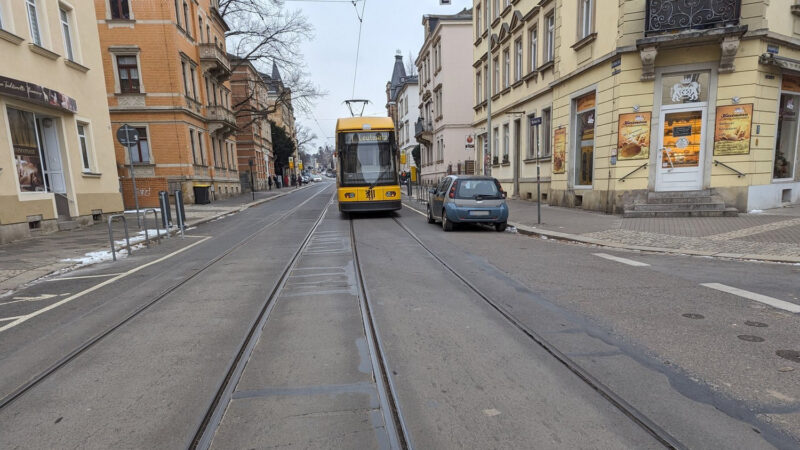 Eine Straße mit zwei Straßenbahngleisen. Auf dem rechten Gleis fährt eine gelbe Straßenbahn. Rechts davon ein kleines Auto am Straßenrand. Hinter dem Auto sieht man die Fahrradbügel. Auf der linken Straßenseite sind ebenso zwei Fahrradbügel.