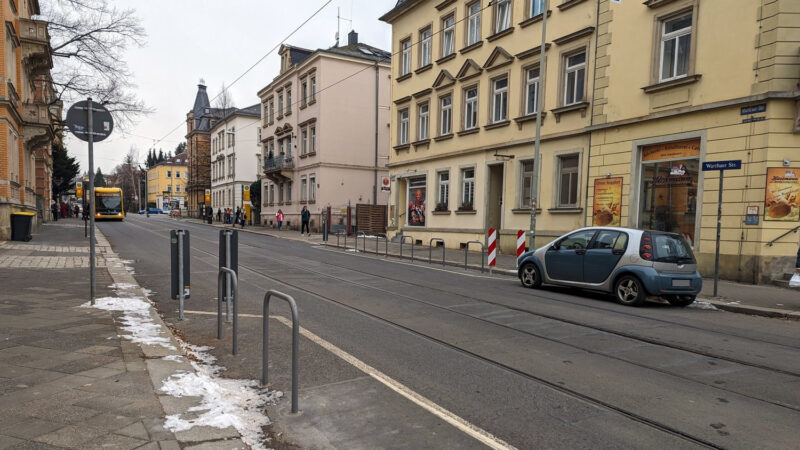 Leere Straße mit zwei Straßenbahngleisen. Im Vordergrund (linke Seite) zwei Fahrradbügel, auf der rechten Seite ein Auto und dahinter 6 Fahradbügel, längs zur Fahtrichtung.