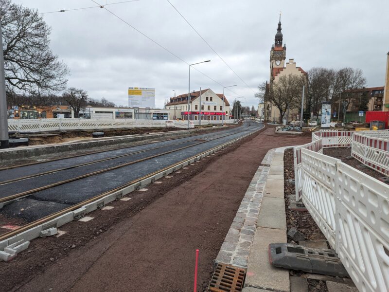 Fast fertige Trasse vor dem Rathaus Cotta, Gleise liegen, dazwischen ist geteert, rechts davon ist noch Sand. Das ganze Feld ist mit weißen Baustellenabsperrungen gesichert.