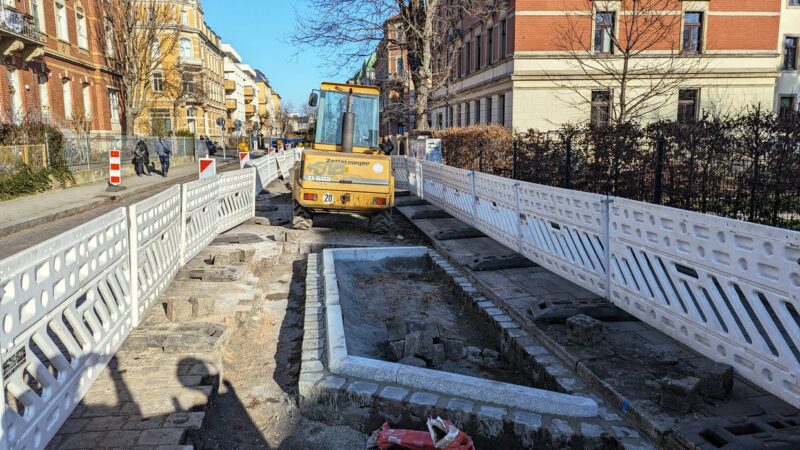 Ein gelber Kleinbagger steht zwischen weißen Absperr-Barken. Davor ein trapezförmiges Feld, welches mit Steinen eingefasst ist. Hier wird später der Baum stehen.