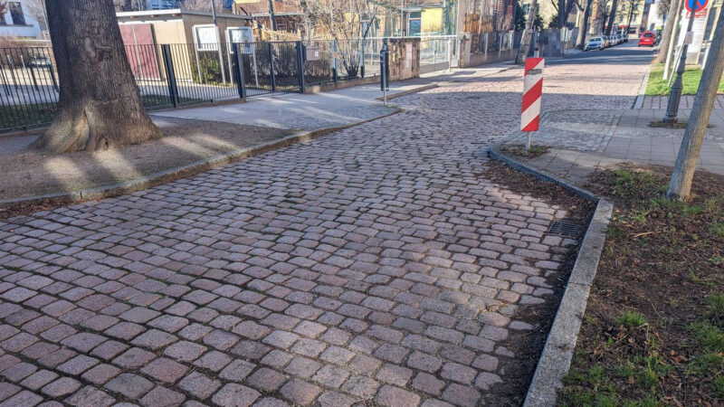 Eine rot-weiße Absperr-Markierung verdeutlicht den vorgezogenen Fußgängerüberweg. Davon und dahinter verbreitert sich die Kopfsteinpflasterstraße. Es sind nur im Hintergrund parkende Autos links und rechts zu sehen.