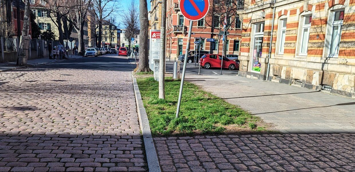 Eine leere Straße, rechts ein Parkverbotsschild, zentral im Hintergrund ein Apothekenschild, rechts ein Haus mit Sandstein und roten Klinkern.