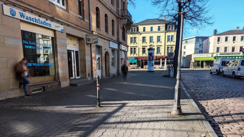 Ein sehr breiter, leerer Gehweg mit einem Poller und einem Leuchten-Mast, rechts eine Kopfsteinpflasterstraße. Im Hintergrund die Häuser der Kesselsdorfer Straße auf die die Bünaustraße hier trifft.