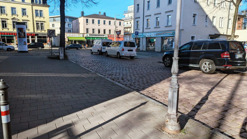 Ein sehr breiter, leerer Gehweg, dann eine Pflasterstraße auf der rechts drei große Autos parken.