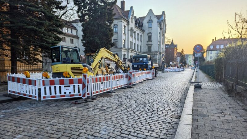 Zwischen rot-weiß markierten Absperrbarken stehen Kleinbagger und ein blauer LKW. Es sind zwei Baustellen auf der linken Straßenseite zu sehen. Auf der Kopfsteinpflaster-Straße besteht während der Bauzeit Halteverbot.
