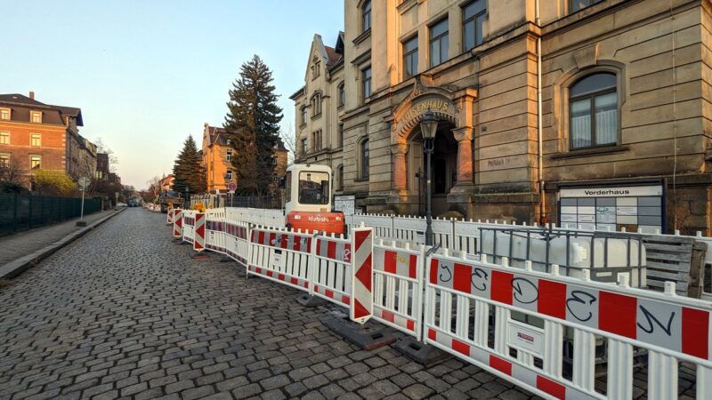 Zwischen rot-weiß markierten Absperrbarken steht ein rot-weißer Kleinbagger vor einem historischen Haus mit der Aufschrift "Lousienhaus". Links davon ist die Kopfsteinpflaster-Straße leer.