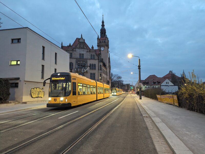 Zwei gelbe Straßenbahnen stehen vor einem historischen Gebäude. Auf der vorderen Bahn steht "Dienstfahrt". Die Straße ist ansonsten leer, frisch geteert, rechts stehen noch Baustellenabsperrungen. Es ist Abend, die Straßenlaternen und die Bahnen leuchten.