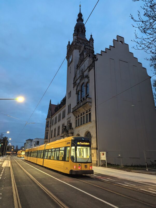 Zwei gelbe Straßenbahnen stehen vor einem historischen Gebäude. Auf der Bahn leuchtet "Testfahrt". Die Straße ist ansonsten leer und frisch geteert. Es ist Abend, die Straßenlaternen leuchten.