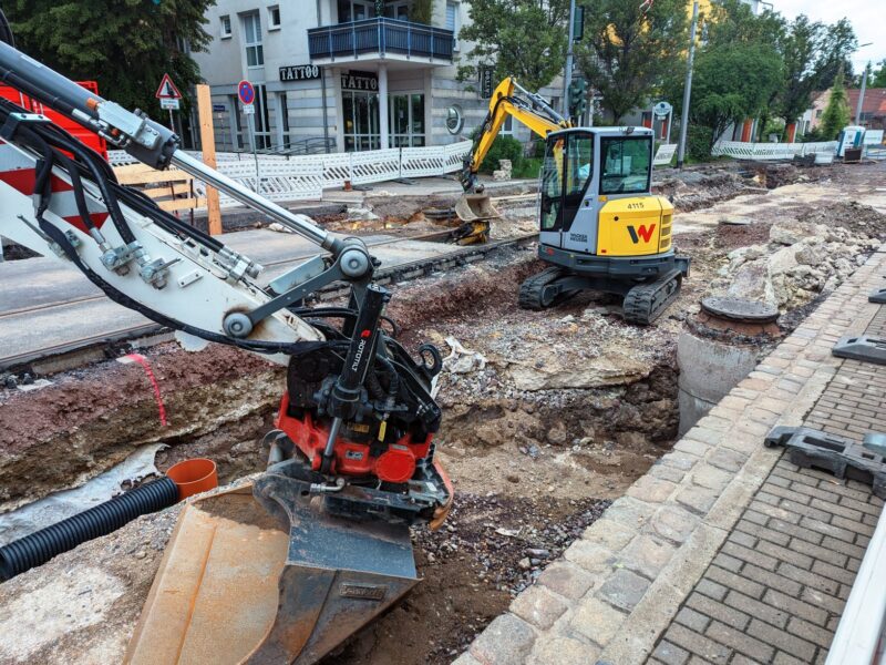 Zwei Bagger stehen in der aufgegrabenen Straße. Links sieht man Reste der Gleise.