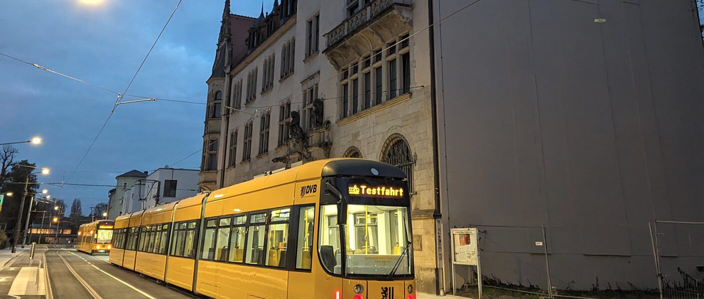 Zwei gelbe Straßenbahnen stehen vor einem historischen Gebäude. Auf der hinteren Bahn steht "Testfahrt". Die Straße ist ansonsten leer, frisch geteert, rechts stehen noch Baustellenabsperrungen. Es ist Abend, die Straßenlaternen und die Bahnen leuchten.