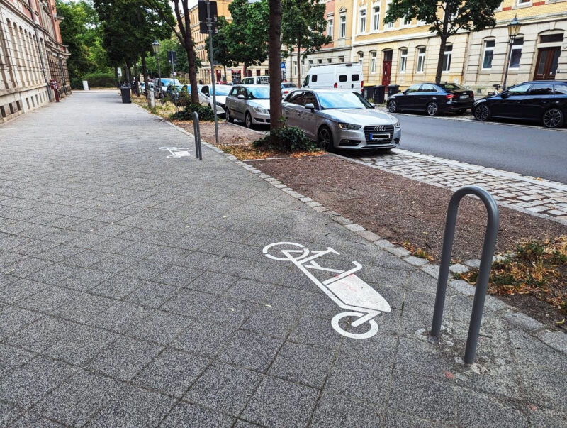 Zwei Lastenrad-Bügel auf dem überbreiten Gehweg. Die Bügel sind mit großen Pktogrammen auf dem Weg gekennzeichnet.