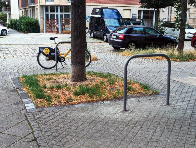 Zwei Fahrradbügel auf dem Gehweg. Einer vor und einer hinter einem Baum an der Kreuzung Bünaustraße / Leumerstraße.