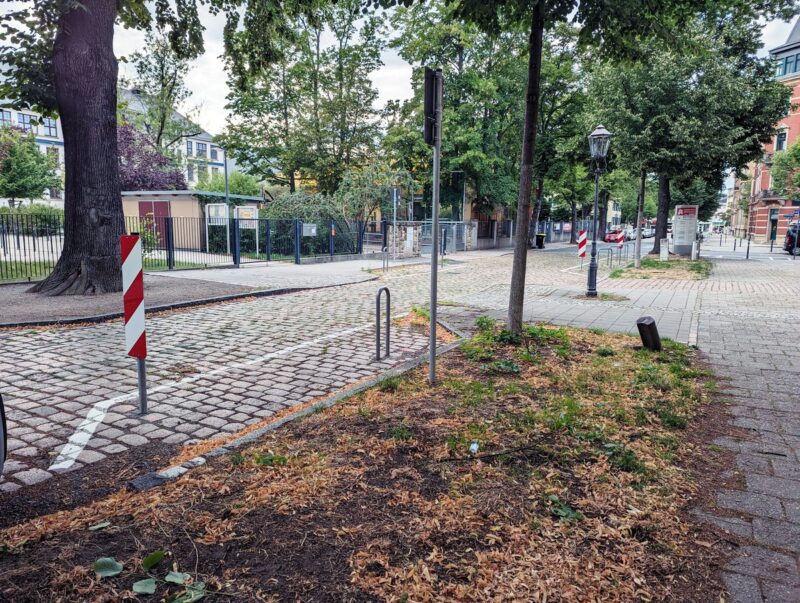 Links vor und rechts hinter einem vorgezogenen Fußgängerüberweg sind Flächen für einen Lastenrad-Bügel abmarkiert. Davor steht eine feste Warnbarke. Pro Fahrradbügel-Standort ist ein Parkplatz entfallen.