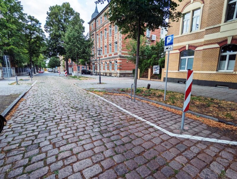 Links vor und rechts hinter einem vorgezogenen Fußgängerüberweg sind Flächen für einen Lastenrad-Bügel abmarkiert. Davor steht jeweils eine feste Warnbarke. Pro Fahrradbügel-Standort ist ein Parkplatz entfallen. Ein Parkplatz-Schild kennzeichnet die Nutzung. Darunter ist ein Lastenrad abgebildet.