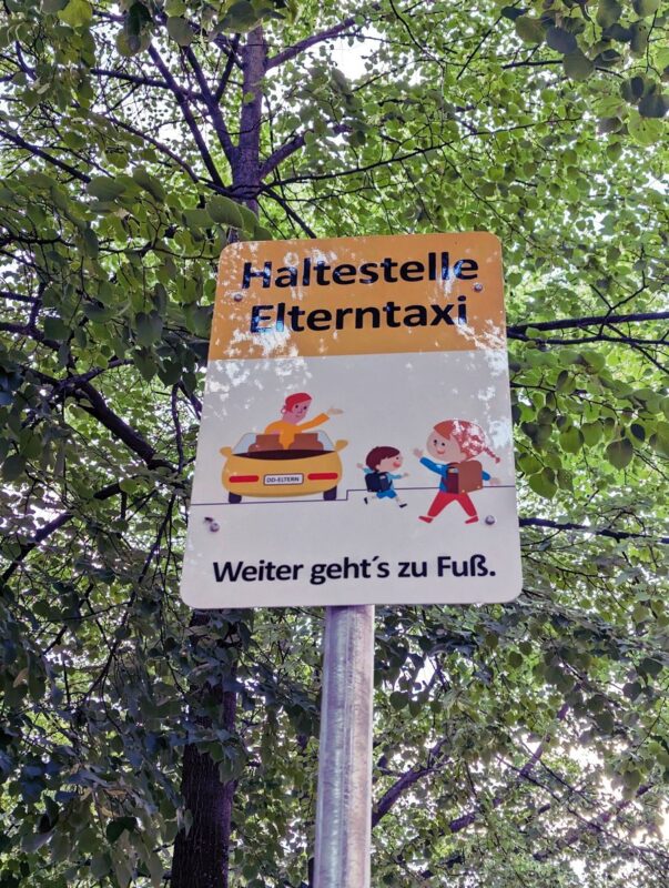 Schild mit Text "Haltestelle Elterntaxi", darunter ein Comic mit einem Auto und zwei Kindern, die sich verabschieden. Unten der Text "Weiter geht's zu Fuß."