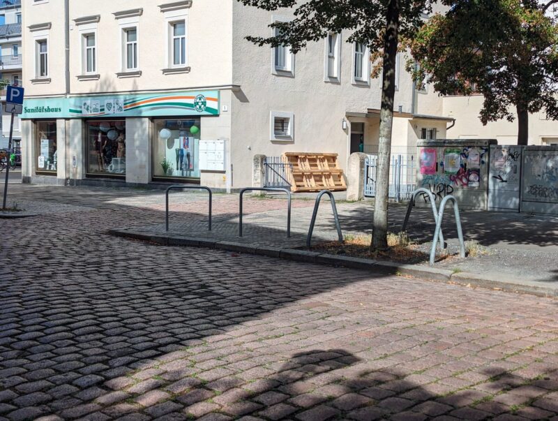 Zwei Fahrradbügel stehen neben einem Baum mit drei Baumschutz-Bügel. Es ist kein Fahrrad angeschlossen.