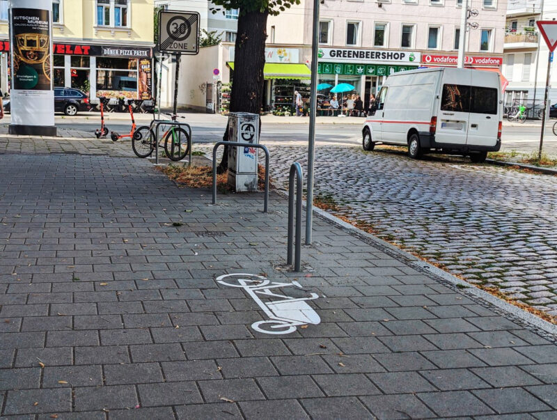 Drei neue Fahrradbügel und ein Lastenradbügel stehen links von der Straße. Am hinteren Fahrradbügel ist ein Fahrrad angeschlossen. Vor dem Lastenradbügel ist ein Piktogram auf den Fußweg gemalt.
