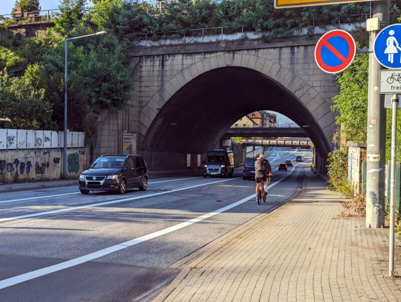 Straße mit mehreren Spuren (Bus-Spur, zwei Fahrspuren, ein Radweg), daneben ein Fußweg, freigegeben für Radfahrer. Mehrere Autos sind auf dem Bild und ein Fahrrad.