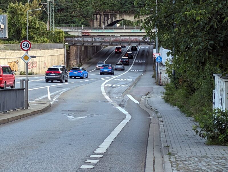 Straße mit mehreren Spuren (Bus-Spur, zwei Fahrspuren, ein Radweg), daneben ein Fußweg, freigegeben für Radfahrer. Mehrere Autos sind auf dem Bild. Blick stadtwärts.