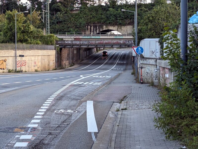 Straße mit mehreren Spuren (Bus-Spur, zwei Fahrspuren, ein Radweg), daneben ein Fußweg, freigegeben für Radfahrer. Ein Autos ist auf dem Bild. Blick stadtwärts.